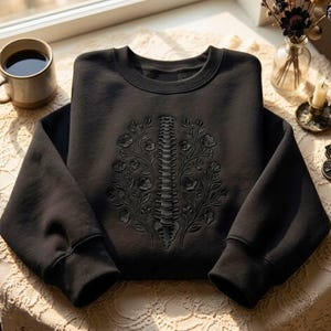 Op de afbeelding: Een zwart sweatshirt met ronde hals en een ton-sur-ton borduursel op de borst. Het ontwerp toont een menselijke ruggengraat in het midden tussen twee symmetrische bloemtakken met bladeren en bloesems, volledig uitgevoerd in zwart garen.