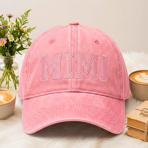 以下が含まれることがあります： 前面に「MIMI」の刺繍が入ったピンク色の野球帽。帽子はカーブしたつばと布製のストラップ留めが付いています。白い表面に、コーヒーカップと小さな花のブーケを背景に表示されています。