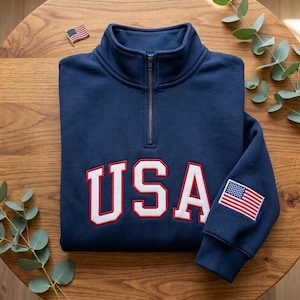 May include: Navy blue pullover sweatshirt with a quarter-zip collar. The word "USA" is embroidered in white with red trim across the chest. An American flag patch is on the left sleeve, and a small flag pin is on the wooden surface.