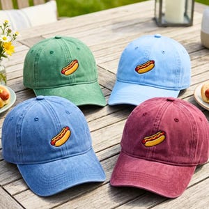 Könnte beinhalten: Vier Baseballkappen in Grün-, Blau- und Burgundertönen liegen auf einem Holztisch. Jede Kappe hat ein gesticktes Hotdog-Design. Die Kappen sind lässig und für den Außenbereich geeignet.