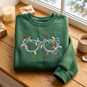 May include: A forest green crewneck sweatshirt featuring an embroidered design of white-rimmed glasses wrapped in colorful Christmas light bulbs in shades of red, blue, yellow, pink, and green, centered on the front of the garment.