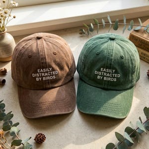 May include: Two baseball caps, one brown and one green, are displayed. Each cap has the phrase "EASILY DISTRACTED BY BIRDS" embroidered in white. The caps are made of a soft, worn fabric, suggesting a casual style. The caps are a great accessory.