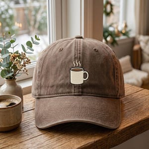 May include: A brown baseball cap featuring an embroidered graphic of a white coffee mug with steam rising from the top. The cap has a structured crown, a curved brim, and a classic six-panel design with stitched eyelets on the top panels.