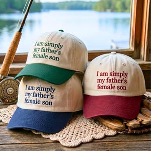 May include: Three baseball caps with cream-colored crowns and colored brims in green, navy blue, and maroon. Each cap features the embroidered text "I am simply my father's female son" in a color matching its respective brim on the front panel.