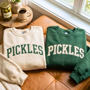 Puede incluir: Dos sudaderas de cuello redondo, una color crema y otra verde oscuro, cada una con la palabra PICKLES en una fuente grande y arqueada de estilo universitario. La sudadera crema tiene letras verde oscuro y la verde oscuro tiene letras crema.