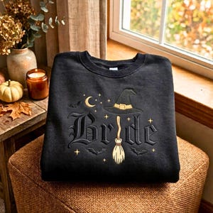 May include: A black crewneck sweatshirt featuring gold embroidered text that reads Bride, with the letter i replaced by a witch's broom. The design includes a witch's hat, a crescent moon, small stars, and flying bats surrounding the lettering.