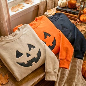Puede incluir: Tres sudaderas de cuello redondo en beige, naranja y negro, cada una con una gran aplicación de cara de calabaza de color oscuro centrada en la parte delantera. Las sudaderas tienen mangas largas y puños acanalados, dispuestas en pila.