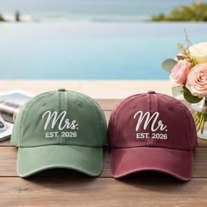 May include: Two baseball caps, one green and one burgundy, sit side-by-side. The green cap says "Mrs. EST. 2026" in white script, and the burgundy cap says "Mr. EST. 2026". A bouquet of flowers is in the background.