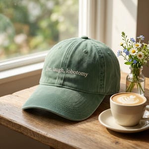 May include: A sage green baseball cap with the text "live, laugh, lobotomy" in white. The cap sits on a wooden table next to a cup of coffee and a small vase of flowers. The hat has a curved brim.