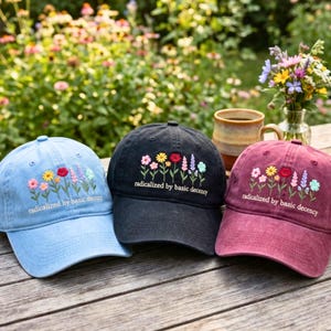 May include: Three baseball caps in light blue, black, and maroon, each featuring embroidered colorful wildflowers and the text "radicalized by basic decency" in white lettering. The caps are arranged side by side on a wooden surface.