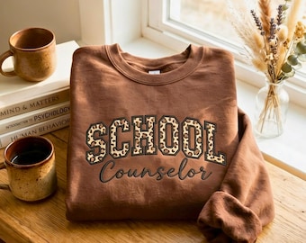 Könnte beinhalten: Ein braunes Sweatshirt mit Rundhalsausschnitt, das den Schriftzug SCHOOL in Blockbuchstaben mit Leopardenmuster und darunter das Wort Counselor in einer schwarzen Schreibschrift zeigt. Das Kleidungsstück hat lange Ärmel sowie gerippte Bündchen.