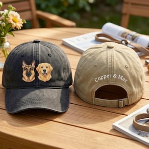 May include: Two baseball caps on a wooden table. One is black with embroidered dog portraits, a German Shepherd and a Golden Retriever. The other is tan with "Copper & Max" embroidered on the back. Both have adjustable straps.