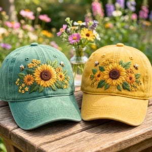 May include: Two baseball caps, one sage green and one mustard yellow, each featuring detailed embroidery of a large central sunflower surrounded by smaller yellow flowers, green leaves, and two small bees on the front panels.