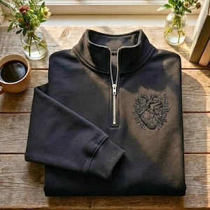 Peut inclure: Un sweat-shirt noir à fermeture éclair quart de zip avec un motif brodé de cœur et de fleurs sur la poitrine. Le sweat-shirt a un col montant et une fermeture éclair argentée. Le vêtement est plié sur une surface en bois.