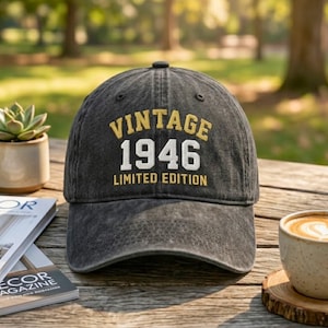 May include: A dark gray baseball cap with the text "VINTAGE 1946 LIMITED EDITION" in gold and white lettering. The cap is sitting on a wooden table with a cup of coffee and a succulent plant in the background.
