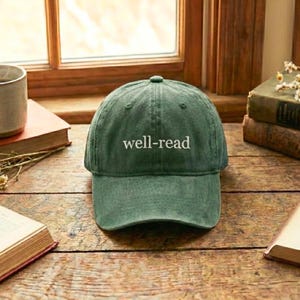 May include: A green baseball cap with the embroidered text "well-read" in white. The hat is sitting on a wooden surface near a window and several books. The cap has a curved brim and a classic six-panel design.