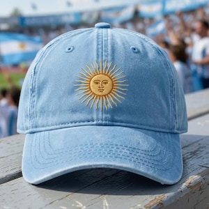 Puede incluir: Una gorra de béisbol de color azul claro con un sol amarillo dorado bordado con un rostro humano en el centro. La gorra tiene una corona estructurada, una visera curva y un botón en la parte superior, con un diseño clásico de seis paneles.