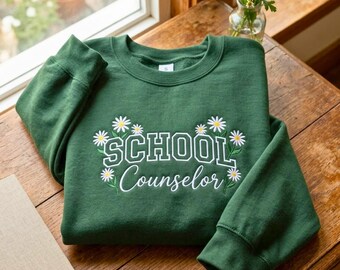 Könnte beinhalten: Ein waldgrünes Sweatshirt mit Rundhalsausschnitt und weißem Sticktext, der SCHOOL Counselor lautet. Das Design ist mit weißen Gänseblümchen mit gelben Mitten und grünen Stielen verziert, die den Schriftzug auf der Vorderseite umgeben.