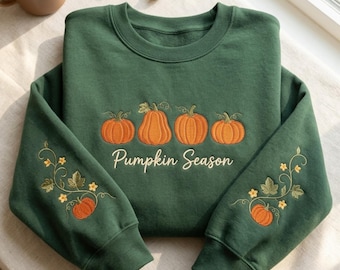 May include: A dark green crewneck sweatshirt featuring an embroidered design of four orange pumpkins across the chest with the text Pumpkin Season in white script. Each sleeve features an embroidered vine with leaves and a small orange pumpkin.