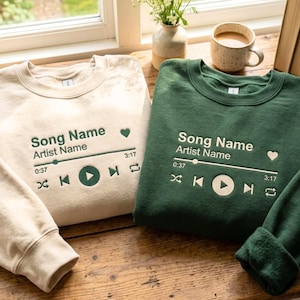Könnte beinhalten: Zwei Sweatshirts mit Rundhalsausschnitt, eines in Creme und eines in Dunkelgrün, mit einem aufgestickten Musikplayer-Grafik auf der Vorderseite. Die Grafik enthält den Text "Song Name" und "Artist Name" sowie Wiedergabe-, Überspring- und Zeitanzeigen.