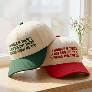 May include: Two baseball caps with the text "I WONDER IF THERE'S A HOT DOG OUT THERE THINKING ABOUT ME TOO". One cap has a green brim and button, the other has a red brim and button. Both caps are beige.