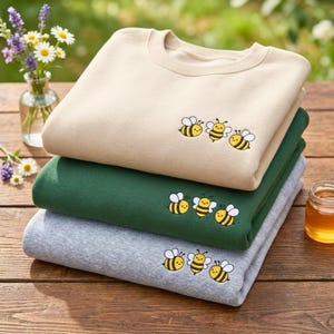 May include: A stack of three crewneck sweatshirts in beige, dark green, and heather gray. Each sweatshirt features an embroidered graphic of three small, smiling cartoon bees with yellow and black striped bodies and white wings on the chest.