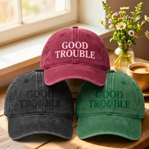 May include: Three baseball caps in burgundy, black, and green, each embroidered with the words "GOOD TROUBLE". The caps are displayed on a wooden surface near a window, with a vase of flowers and a cup of coffee in the background.