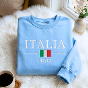 Op de afbeelding: Een lichtblauw sweatshirt met ronde hals voorzien van het woord ITALIA geborduurd in witte hoofdletters boven een kleine rechthoekige Italiaanse vlag en het woord ITALY geborduurd in witte hoofdletters onder de vlag op de borst.