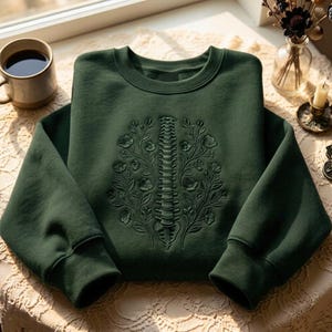 Op de afbeelding: Een donker bosgroene sweater met ronde hals en een ton-sur-ton borduursel op de borst. Het borduursel toont een menselijke ruggengraat in het midden tussen twee symmetrische bloementakken met bladeren en bloesems, uitgevoerd in groen garen.
