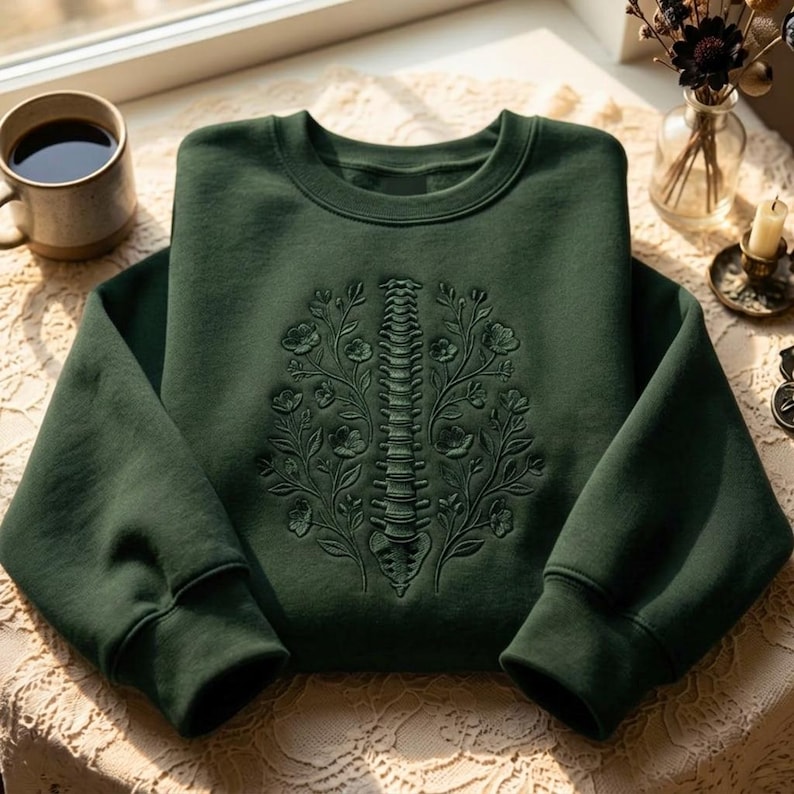 Op de afbeelding: Een donker bosgroene sweater met ronde hals en een ton-sur-ton borduursel op de borst. Het borduursel toont een menselijke ruggengraat in het midden tussen twee symmetrische bloementakken met bladeren en bloesems, uitgevoerd in groen garen.