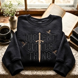 May include: A black long-sleeve sweatshirt featuring gold and black embroidery. The text reads VIOLENT LITTLE THING, with a central gold sword replacing the letter I and three small gold dragon figures surrounding the lettering.