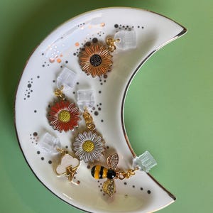 May include: A collection of decorative earrings featuring floral and insect designs, including sunflowers, daisies, and a bee. The earrings are displayed on a crescent-shaped, white ceramic dish with gold trim and speckled accents. The background is a solid green.