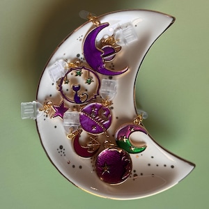 May include: A white crescent-shaped trinket dish with a gold rim, holding several celestial-themed charms. The charms feature moons, stars, and planets in shades of purple, pink, and green. The dish is decorated with small gold dots.
