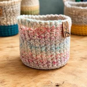 May include: A cylindrical crochet basket crafted from variegated yarn in shades of cream, soft pink, and light blue. The basket features a small, rectangular brown leather tag attached to the rim, embossed with a floral icon and the letters PS.