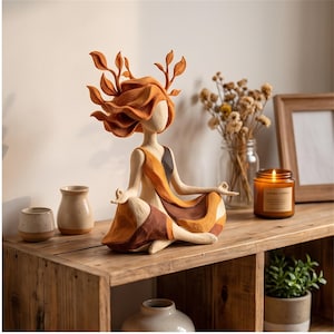 May include: A decorative figurine of a person sitting in a cross-legged meditation pose. The figure has hair styled like orange leaves and branches, wearing a flowing garment in shades of cream, orange, and dark brown with a textured finish.