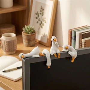 Könnte beinhalten: Vier kleine weiße Gänsefiguren mit orangefarbenen Schnäbeln und Füßen sitzen auf der Oberkante eines Computermonitors. Jede Gans nimmt eine andere Pose ein, darunter liegend, über den Rand hängend, aufrecht sitzend und ruhend.