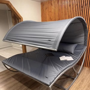 Puede incluir: Una moderna cama balancín gris con estructura metálica curvada y dosel. La cama tiene colchón, almohada y dosel grises. Un portátil y un libro abierto descansan sobre la cama. El dosel tiene el texto "3BA".