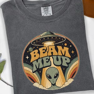May include: Dark gray t-shirt with a retro graphic design featuring a UFO, an alien, and the words "BEAM ME UP." The design includes a rainbow and stars. The shirt is made of a soft material.