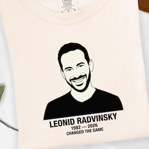 May include: Cream-colored t-shirt featuring a black and white portrait of a smiling man. Below the portrait is the text "LEONID RADVINSKY 1982-2026 CHANGED THE GAME". The shirt is folded and has a Comfort Colors tag.