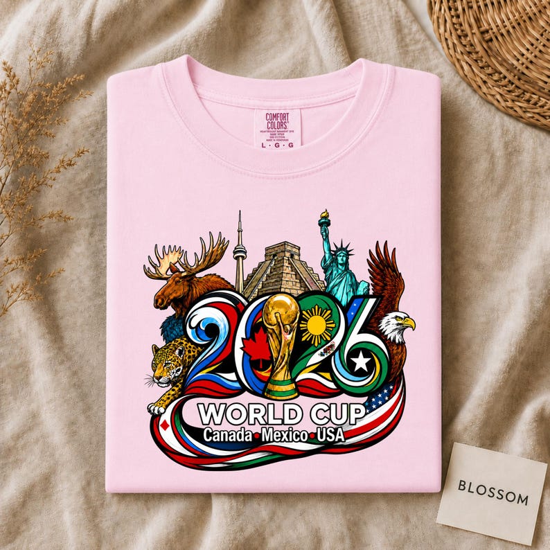 May include: A light pink t-shirt featuring a colorful graphic of the 2026 World Cup. The design includes a moose, a jaguar, the Statue of Liberty, a bald eagle, the CN Tower, a pyramid, and a trophy, with the text 2026 WORLD CUP Canada Mexico USA.
