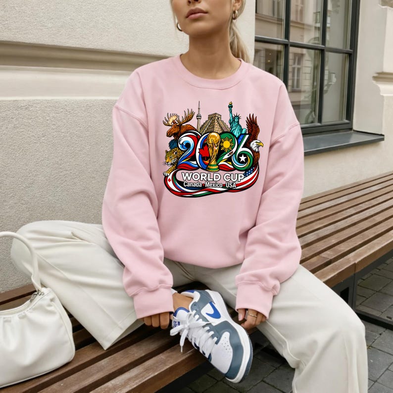 May include: A light pink crewneck sweatshirt featuring a colorful graphic print of the 2026 World Cup logo. The design includes illustrations of a moose, a pyramid, the Statue of Liberty, and an eagle, with the text World Cup Canada Mexico USA.