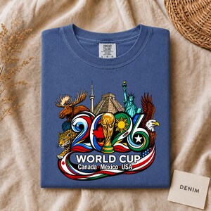 May include: A blue t-shirt featuring a graphic print of the 2026 World Cup logo with the text "WORLD CUP Canada Mexico USA". The design includes illustrations of a moose, a jaguar, the Statue of Liberty, a bald eagle, and a soccer trophy.