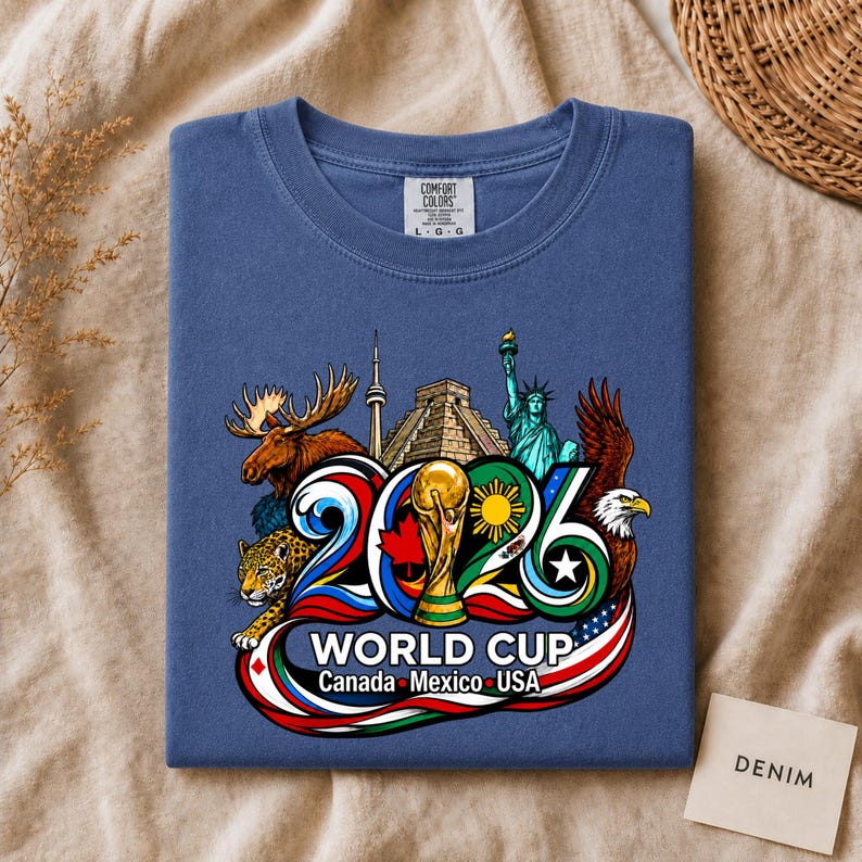 May include: A blue t-shirt featuring a graphic print of the 2026 World Cup logo with the text "WORLD CUP Canada Mexico USA". The design includes illustrations of a moose, a jaguar, the Statue of Liberty, a bald eagle, and a soccer trophy.
