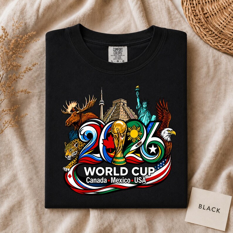 May include: A black t-shirt featuring a colorful graphic of the 2026 World Cup. The design includes a moose, a pyramid, the Statue of Liberty, a jaguar, and a bald eagle, with the text 2026 WORLD CUP Canada Mexico USA printed below.