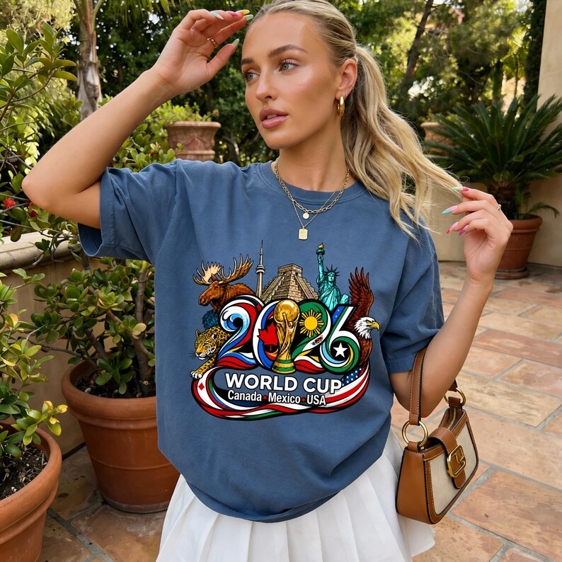 May include: A blue short-sleeve t-shirt featuring a graphic print of the 2026 World Cup logo. The design includes a moose, a jaguar, the Statue of Liberty, an eagle, a pyramid, and the World Cup trophy, with the text Canada Mexico USA.