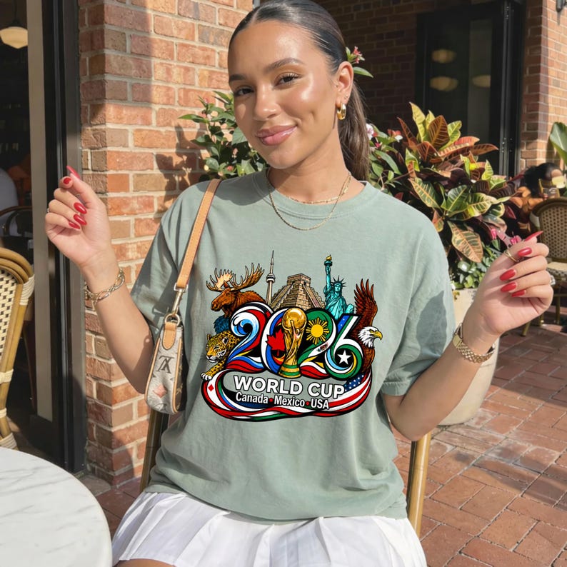 May include: A sage green short-sleeve t-shirt featuring a colorful graphic print of the 2026 World Cup. The design includes a moose, a jaguar, the Statue of Liberty, a bald eagle, and the text 2026 WORLD CUP Canada Mexico USA.