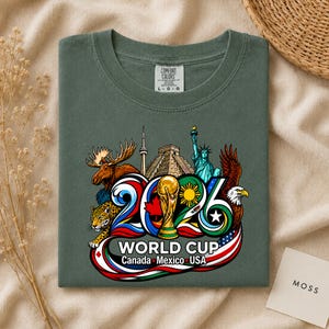 May include: A dark olive green t-shirt featuring a graphic of the 2026 World Cup with the text Canada, Mexico, USA. The design includes a moose, a jaguar, the CN Tower, a pyramid, the Statue of Liberty, a soccer trophy, and a bald eagle.