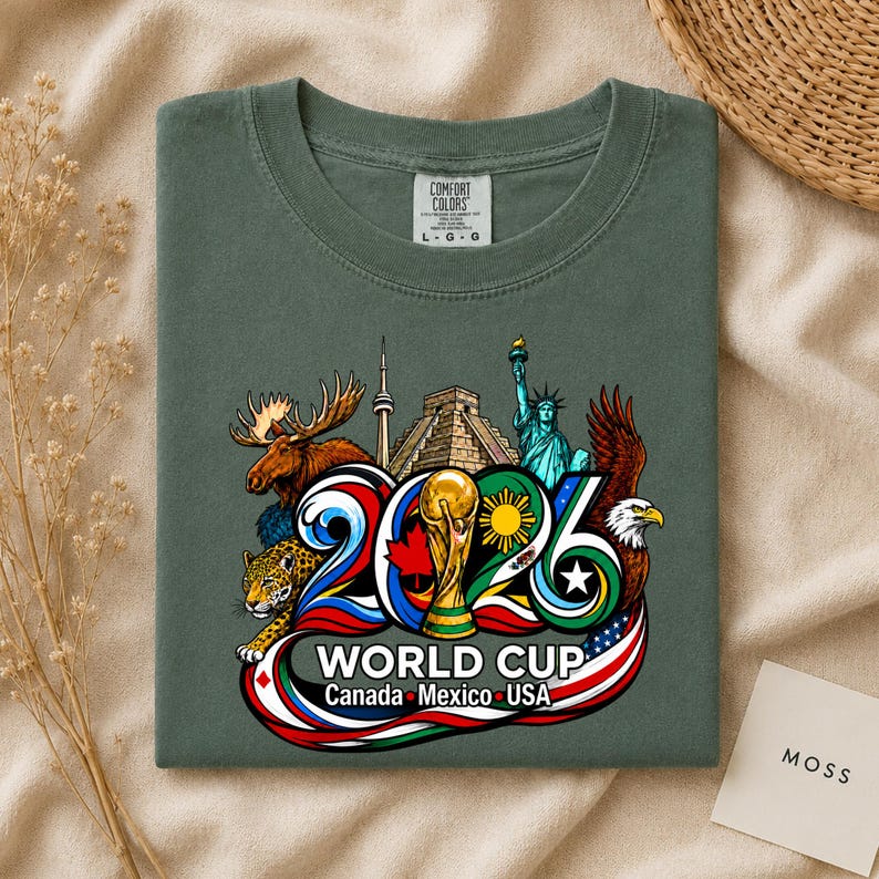 May include: A dark olive green t-shirt featuring a graphic of the 2026 World Cup with the text Canada, Mexico, USA. The design includes a moose, a jaguar, the CN Tower, a pyramid, the Statue of Liberty, a soccer trophy, and a bald eagle.