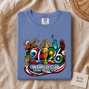 May include: A blue crew neck t-shirt featuring a graphic print of the 2026 World Cup logo. The design includes a moose, a jaguar, the Statue of Liberty, a bald eagle, the CN Tower, a pyramid, and a soccer trophy with the text World Cup Canada Mexico USA.