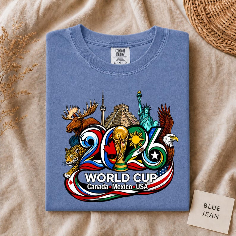May include: A blue crew neck t-shirt featuring a graphic print of the 2026 World Cup logo. The design includes a moose, a jaguar, the Statue of Liberty, a bald eagle, the CN Tower, a pyramid, and a soccer trophy with the text World Cup Canada Mexico USA.